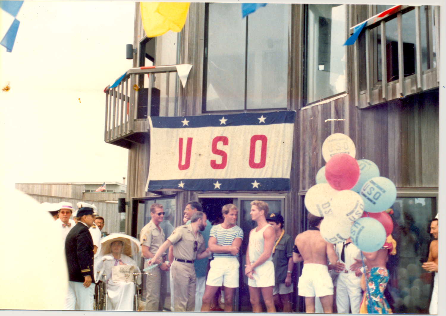 Men in summery clothes outside a building with balloons, bunting and a red, white and blue flag with stars that says USO.