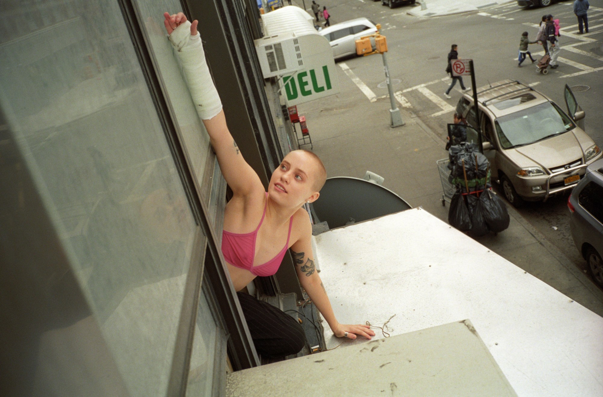 a person in a hot pink triangle bra with a buzzcut and a cast on their arm leans out of a NYC window