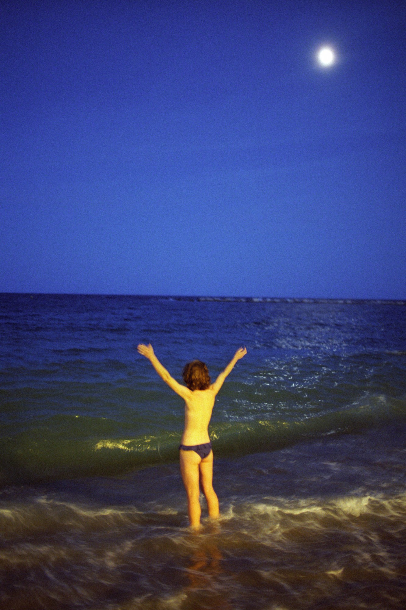 a person stands topless in the sea a night with their arms in the air