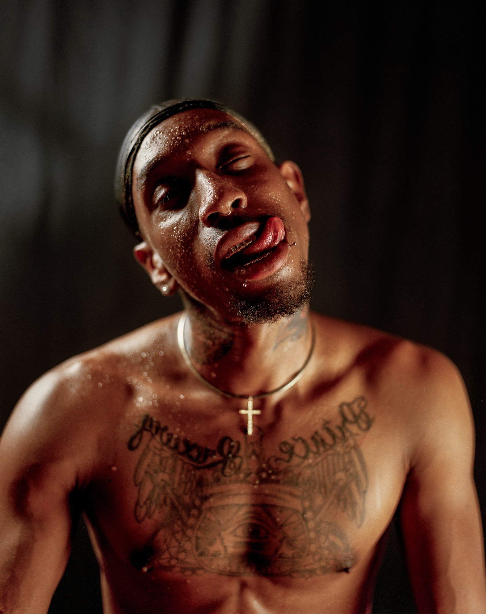 Shirtless man with a crucifix necklace covered in water and sticking his tongue out.