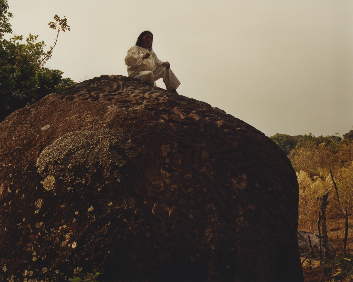 A man sits on top of a large rock in the forest.