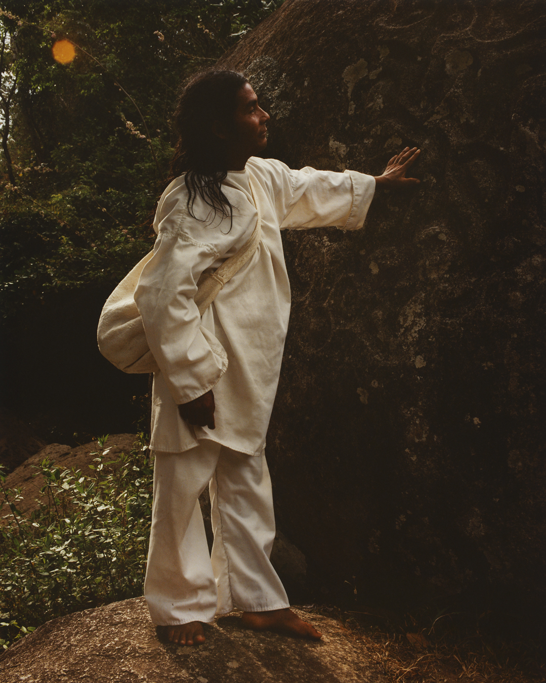 Man in white cotton clothing standing on a rock and touching a larger rock.