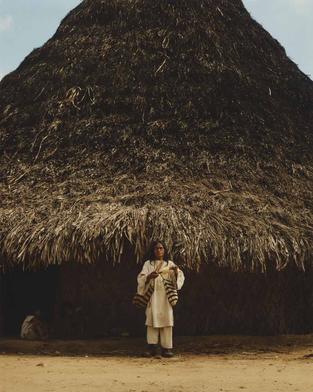 A man in white linens with two stripy bags standing outside a hut with a large funnelled reed roof.