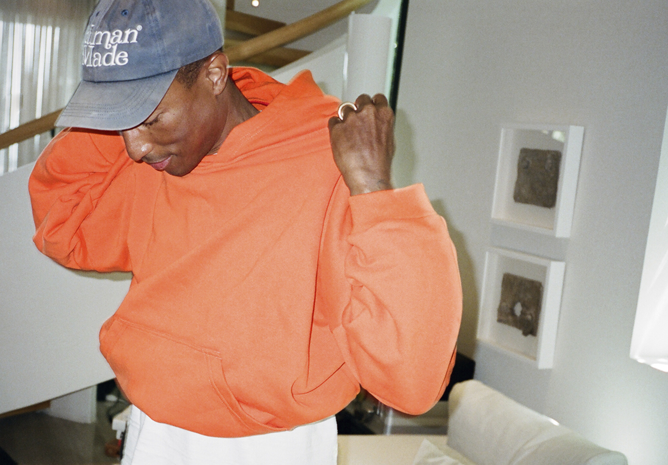 Pharrell Williams wearing an orange hoodie, white trousers and a denim cap