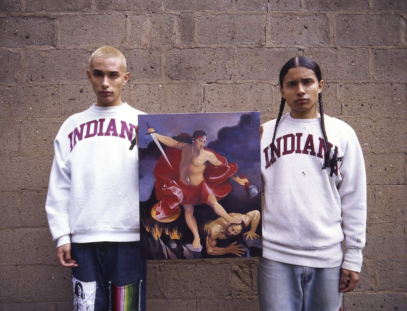 Victor Vegas and model wearing INDIAN sweatshirts and holding a painting of a person standing on another man's head.