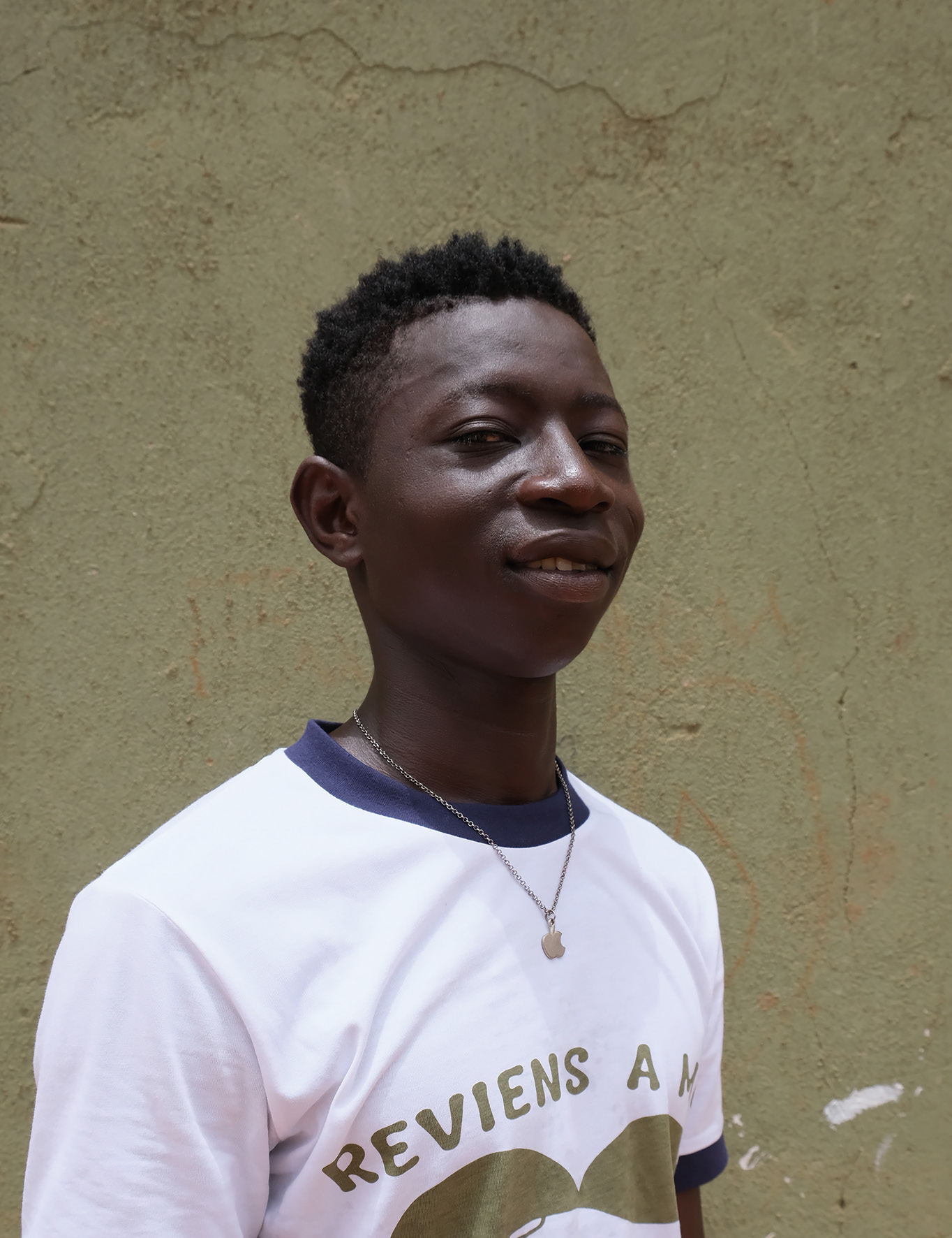 Boy against a wall in a t-shirt and apple necklace.