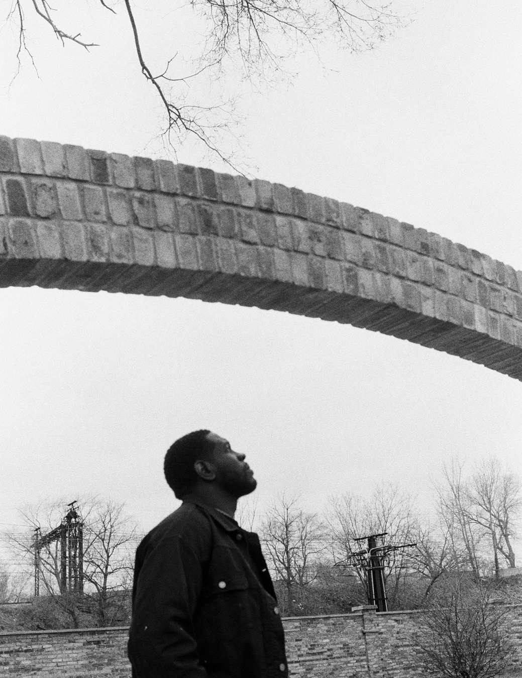 Portrait of Easy Otaber standing under a bridge by a brick wall and power lines.