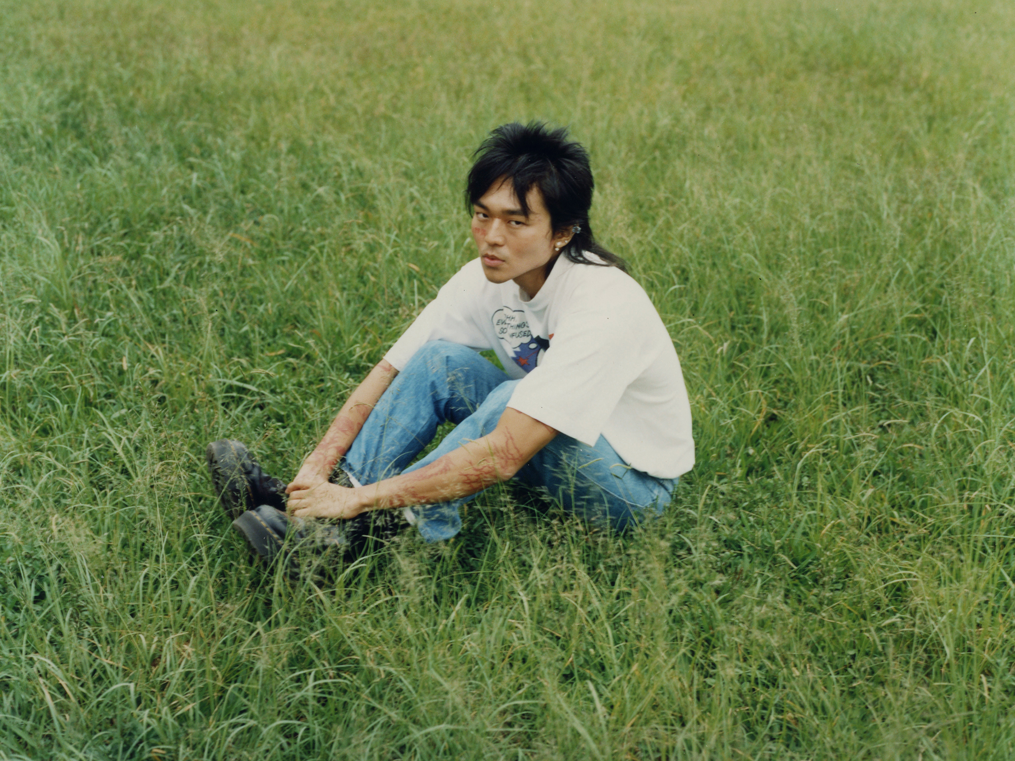 a person with a mullet, wearing jeans and a t-shirt, sits hugging their knees in a field