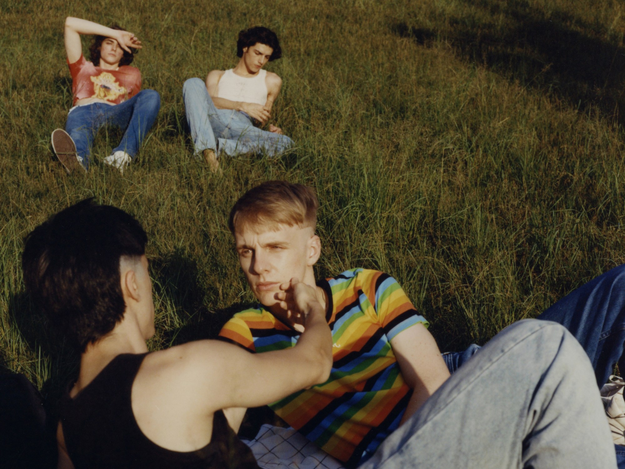 a group of four young men lounge about in a field; one touches another's face tenderly