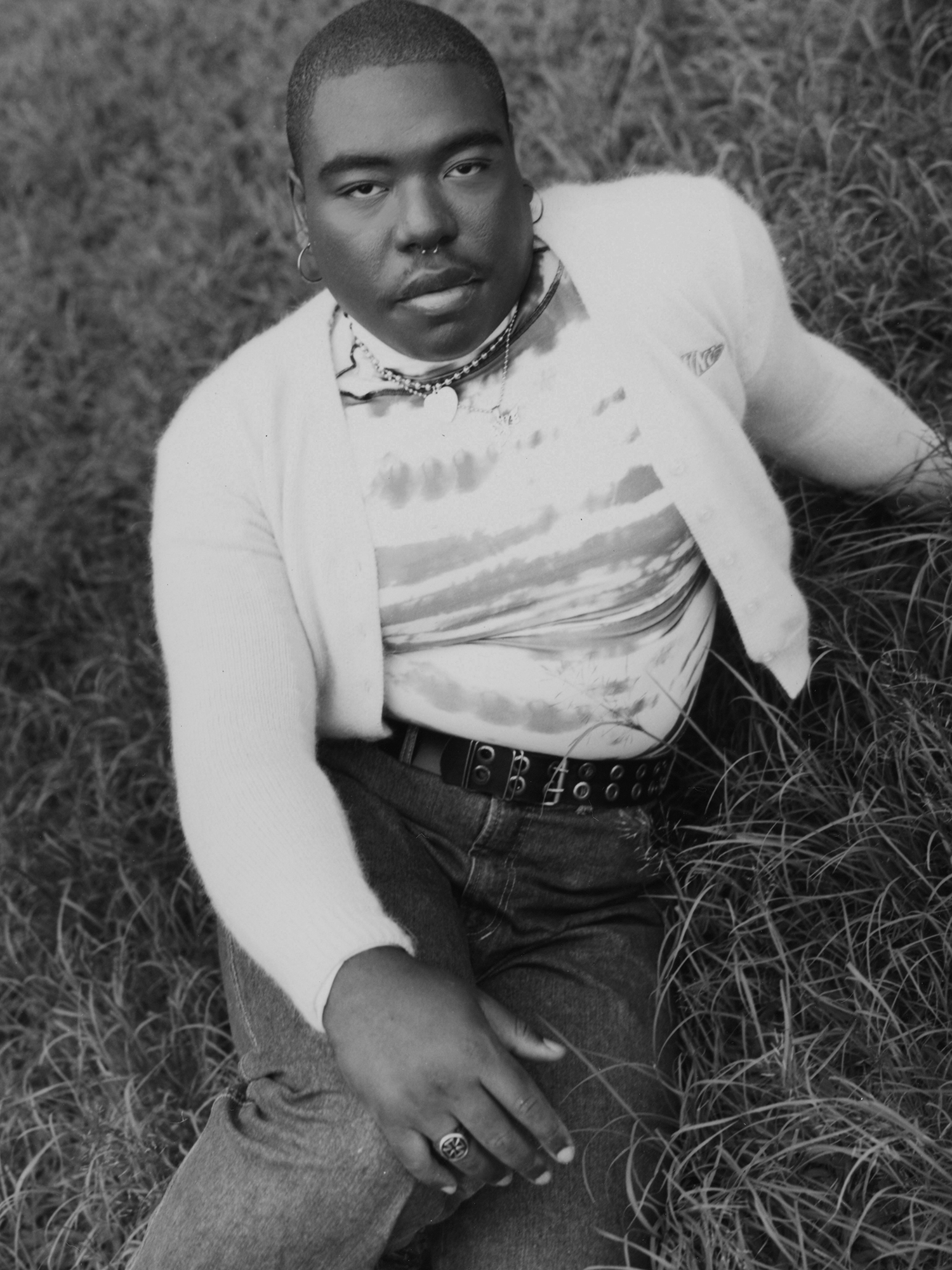 a black and white portrait a man wearing jewellery and a cardigan, sitting in a field