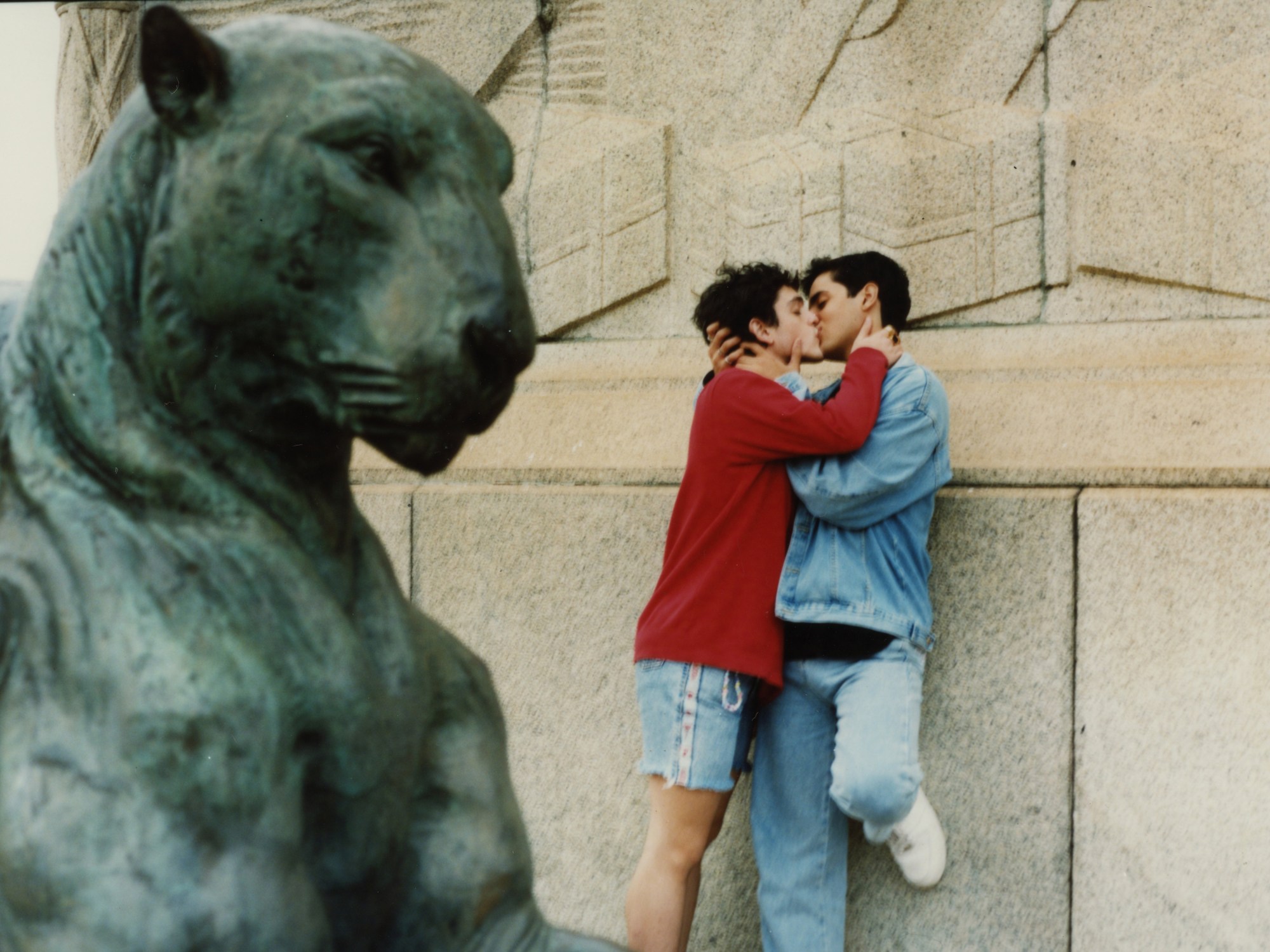 two men embrace against a stone wall; a statue of a lion is in the foreground