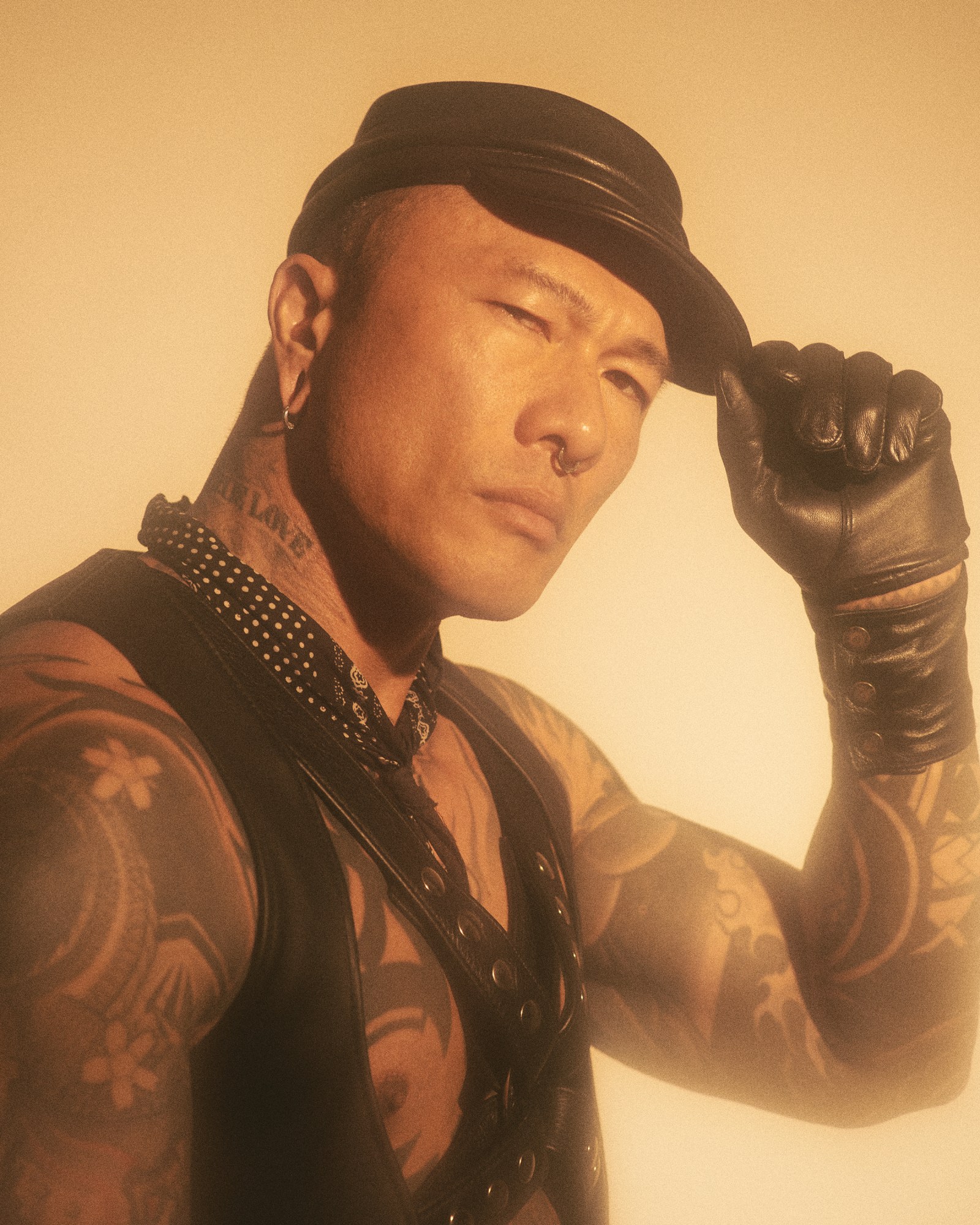 person with tattoos holding their leather hat wearing a leather waistcoat, harness and a bandanna