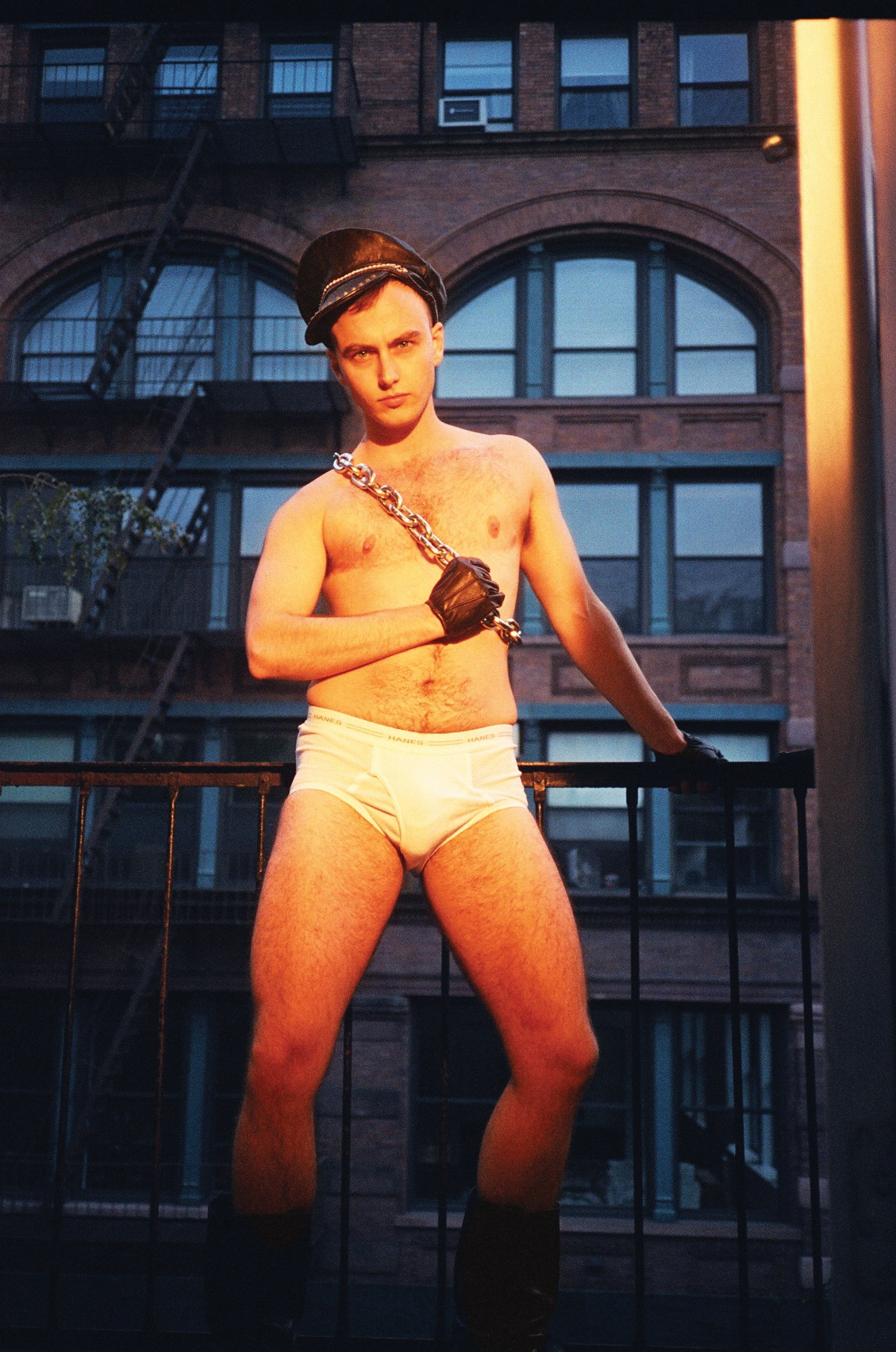 person in white briefs, black boots a chain over their shoulder and leather gloves and hat on standing on an apartment balcony.