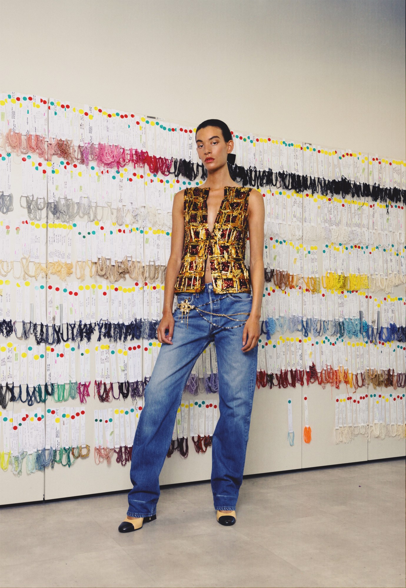 a model wears blue jeans and a golden embroidered top, posing in front of a wall of colourful thread samples