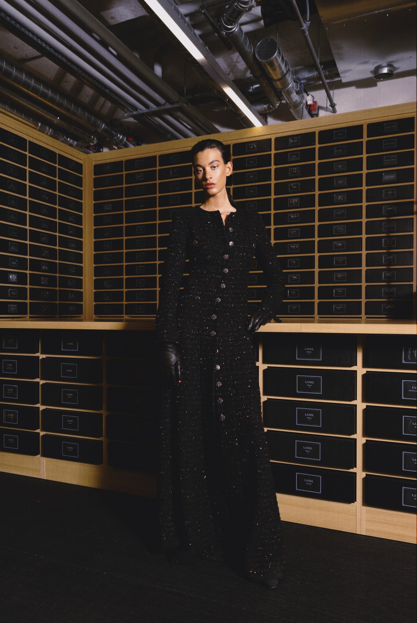 a model wears a long black tweed chanel coat in a dark room of drawers