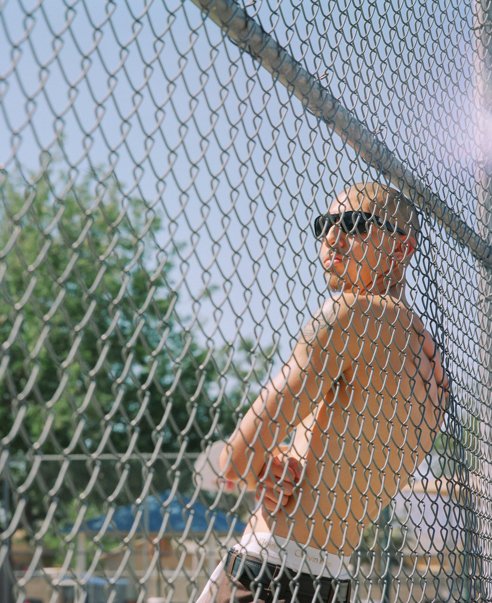 a topless man in sunglasses leaning against a chain fence