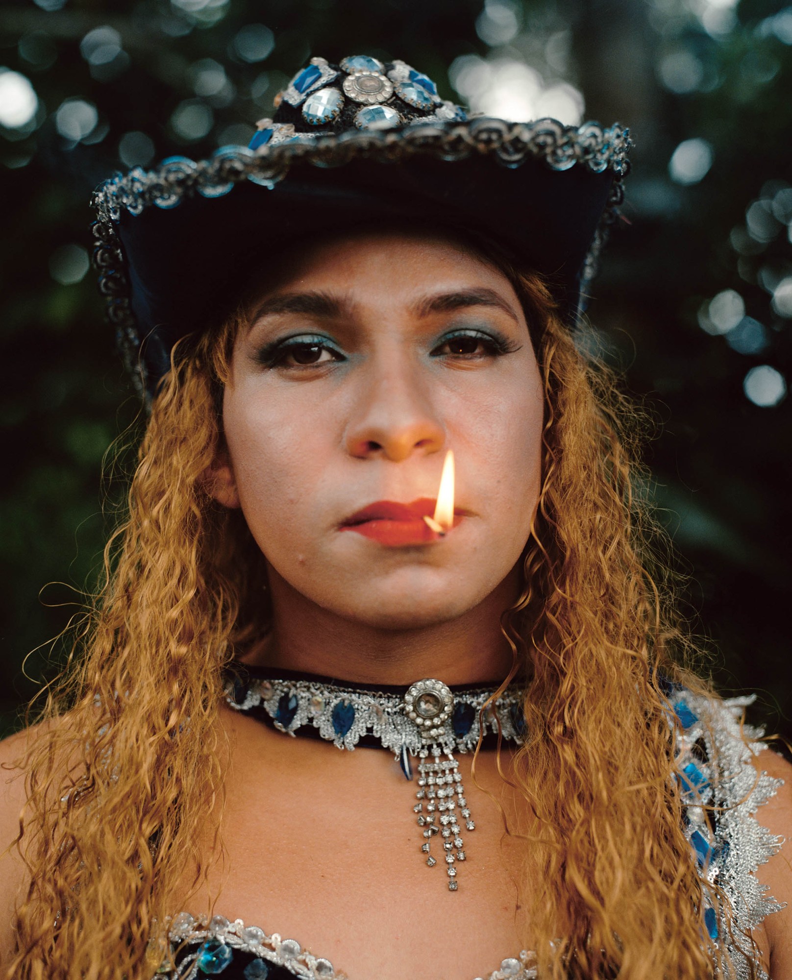 A young person whearing a cowboy hat and choker covered in crystals while holding a lit match in their mouth.