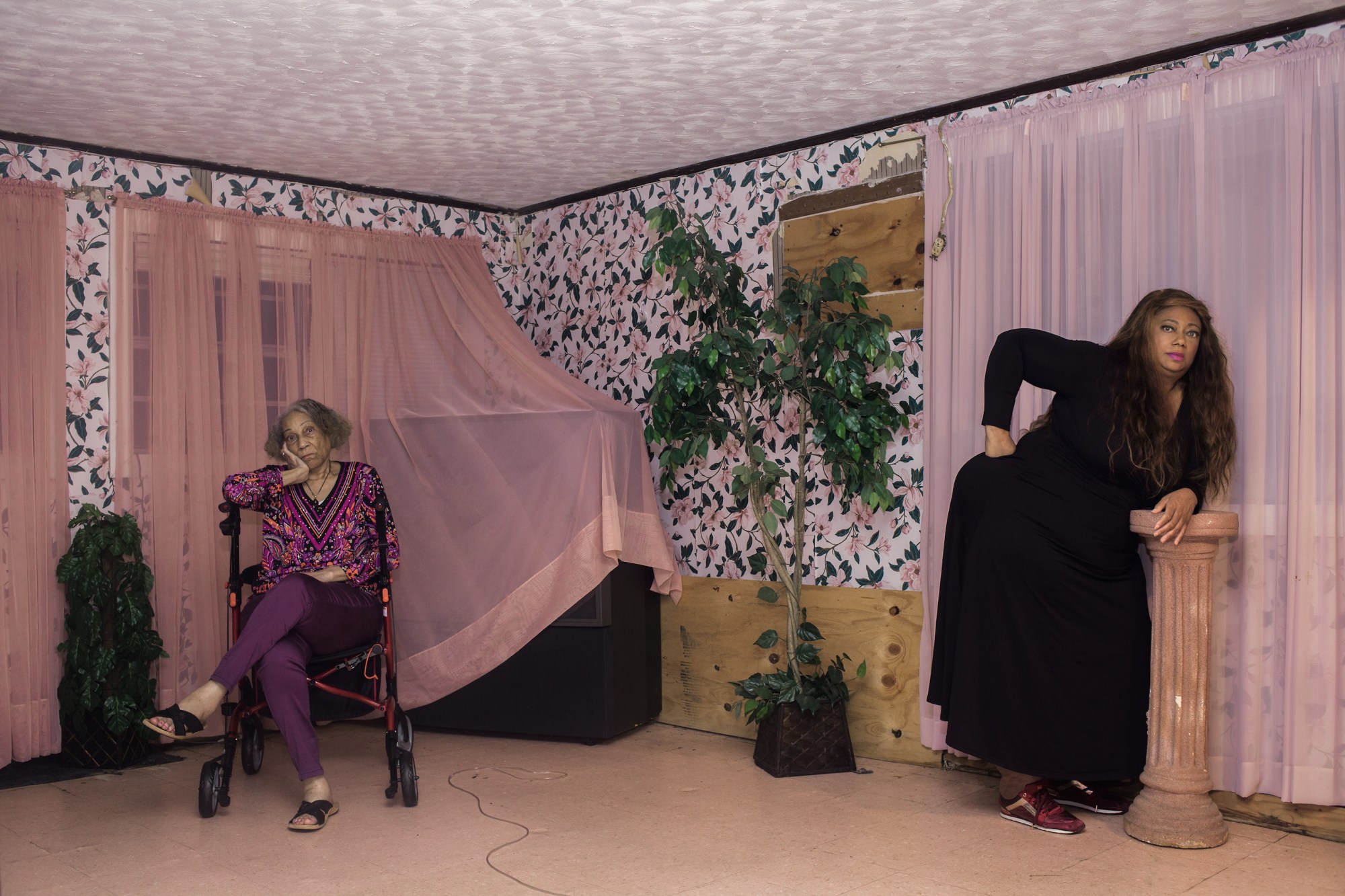 two people in an overly decorated room pose on opposite sides, sitting on a wheelchair and resting on a plinth