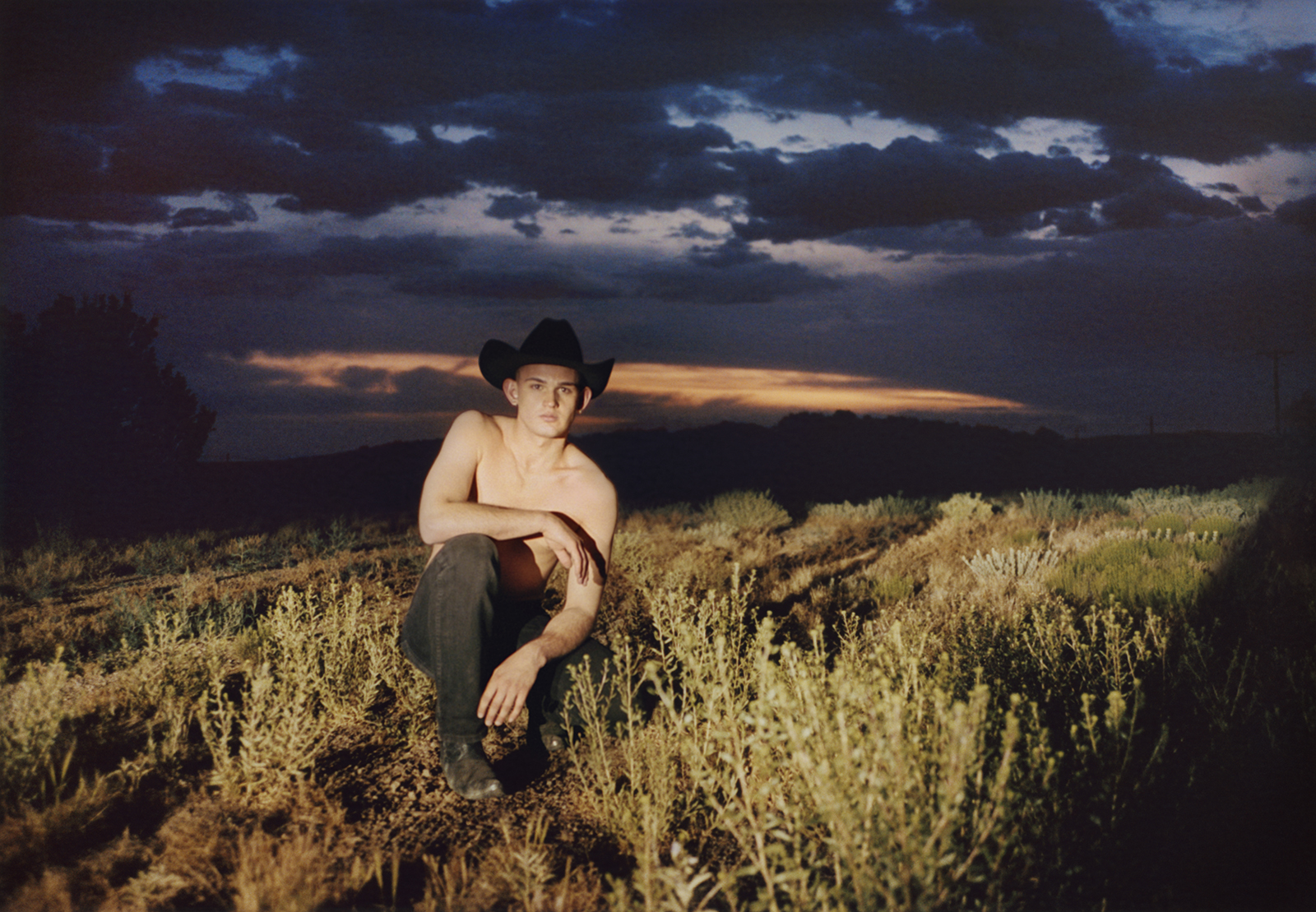 a cowboy crouches in a field at sunset