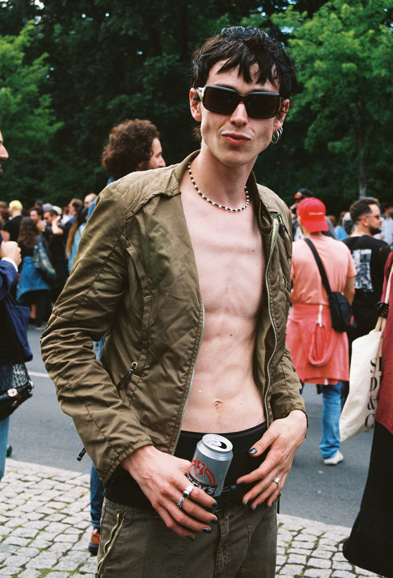 a skinny man holding a beer stands looking to camera, his shirt open