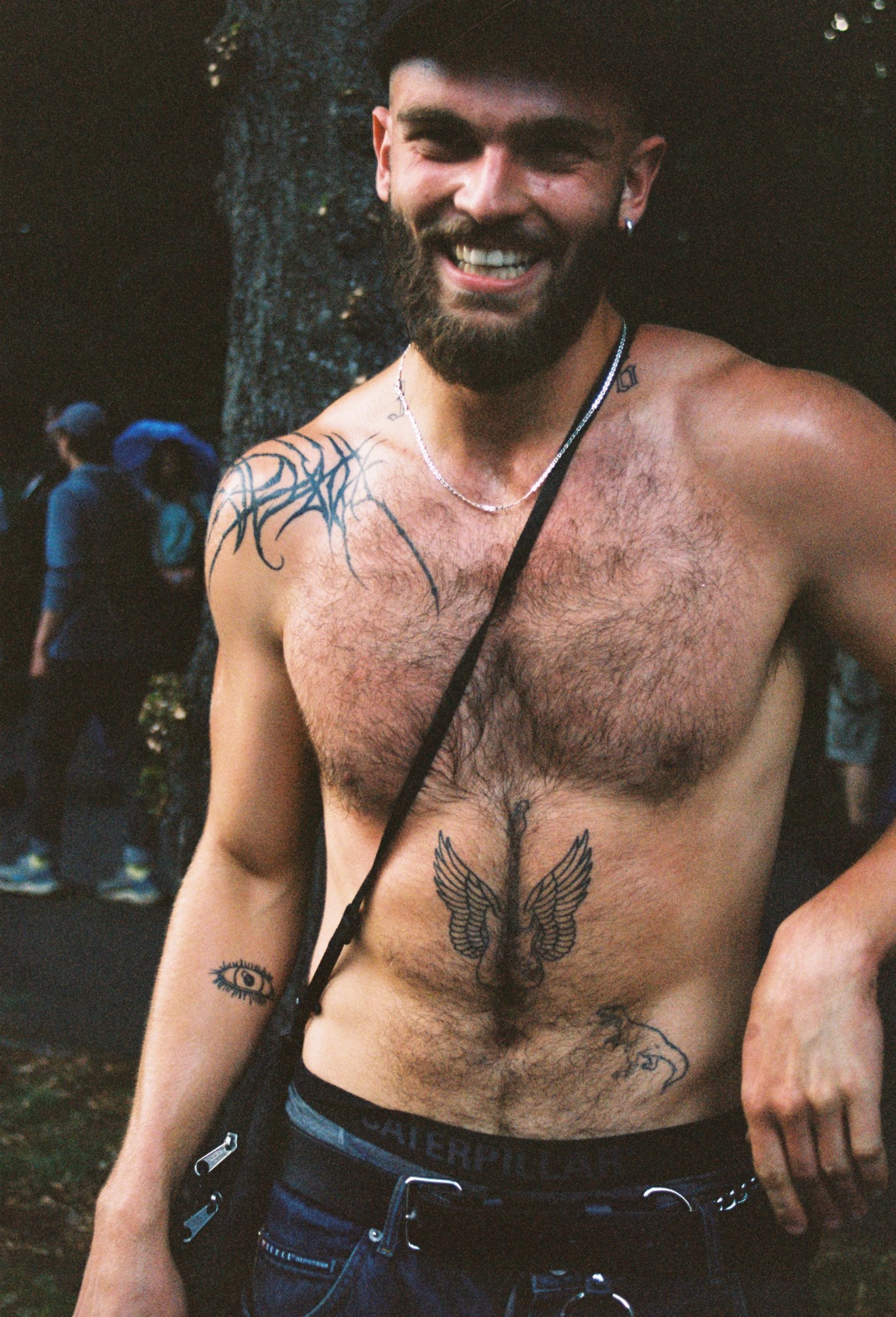 a tatooed man with a hair chest and a beard smiles to the camera