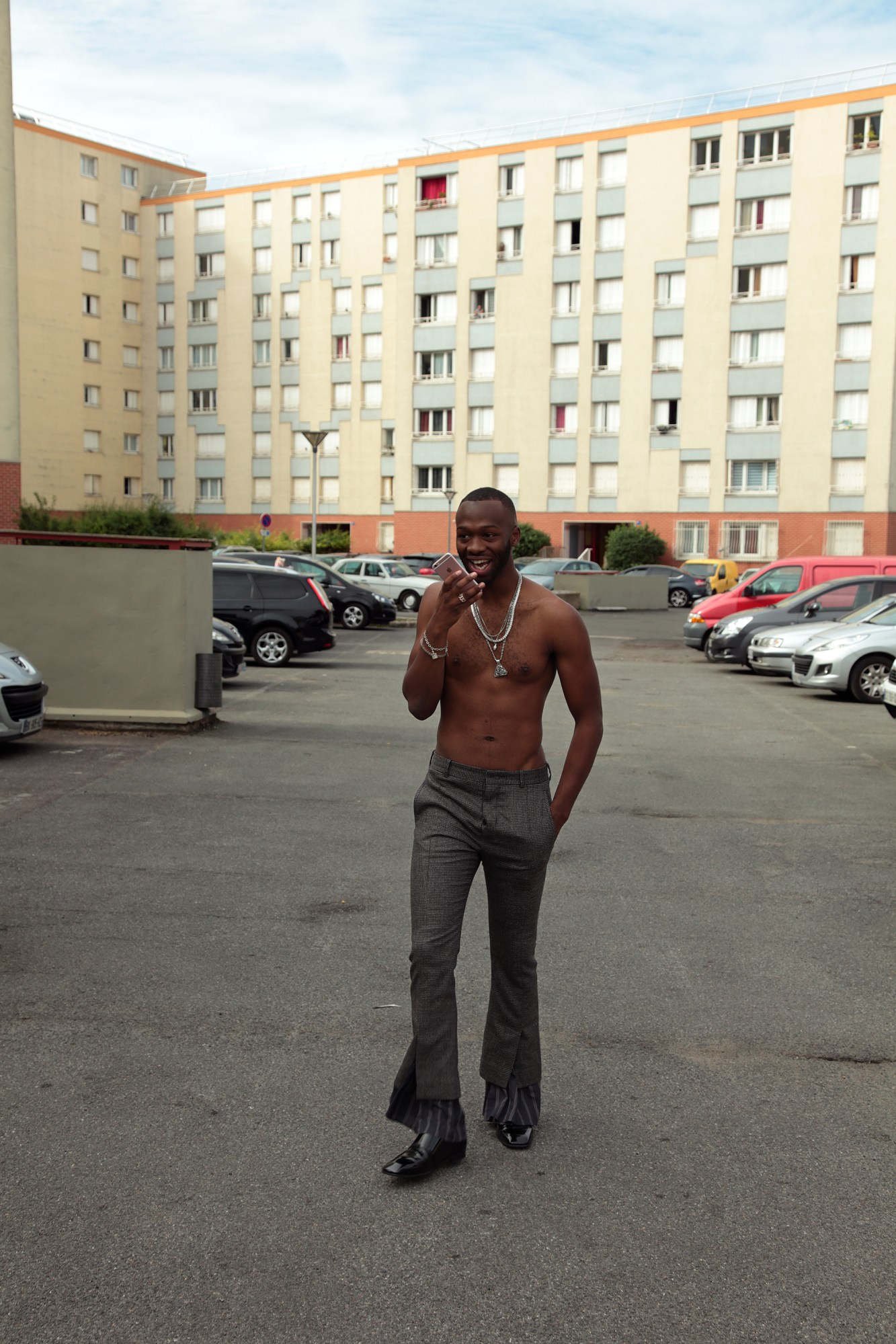 a topless man in grey bootcut pants walks aross a car park talking on a pink iphone