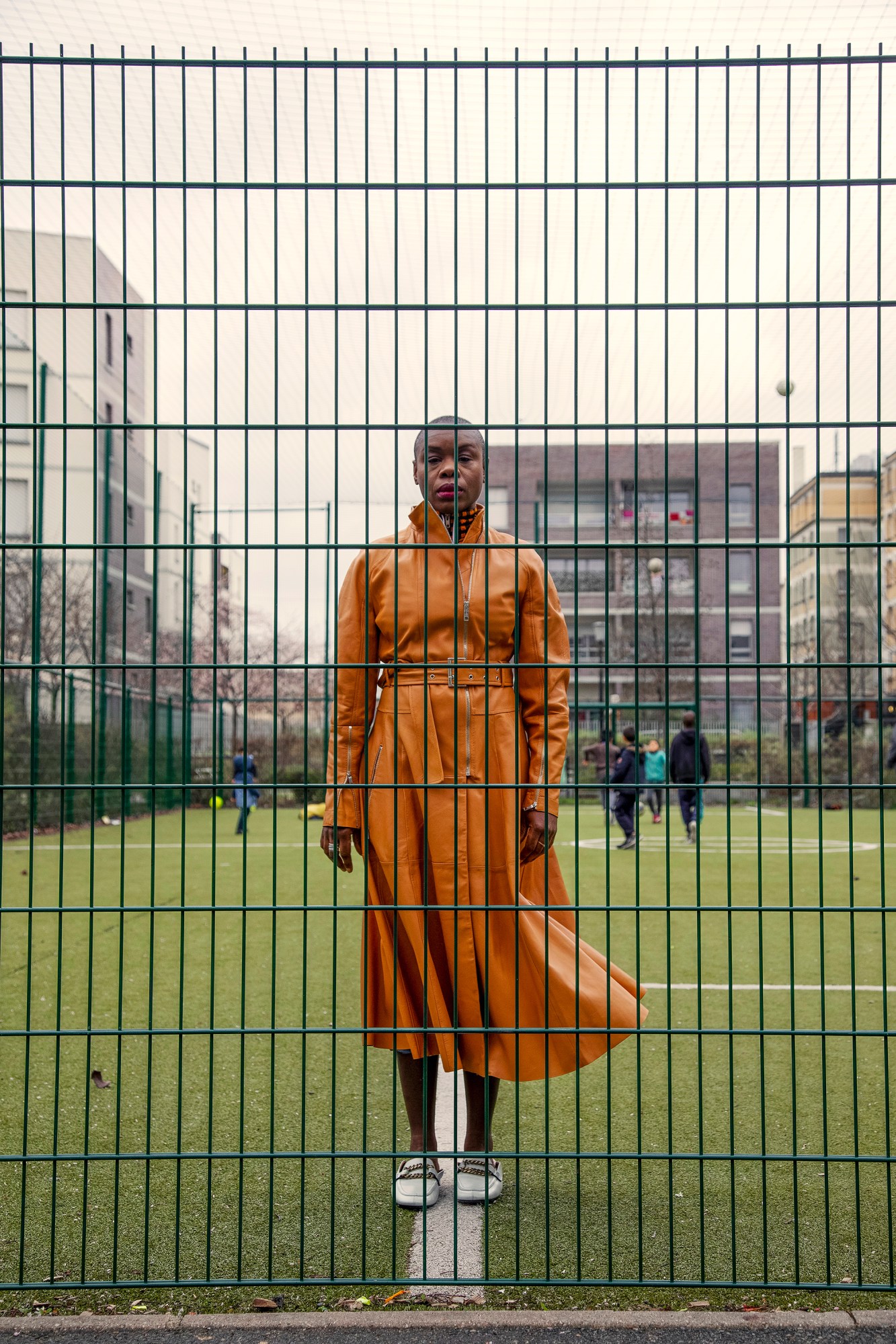 a woman in an orange leather dress stands behind the railings of a football pitch