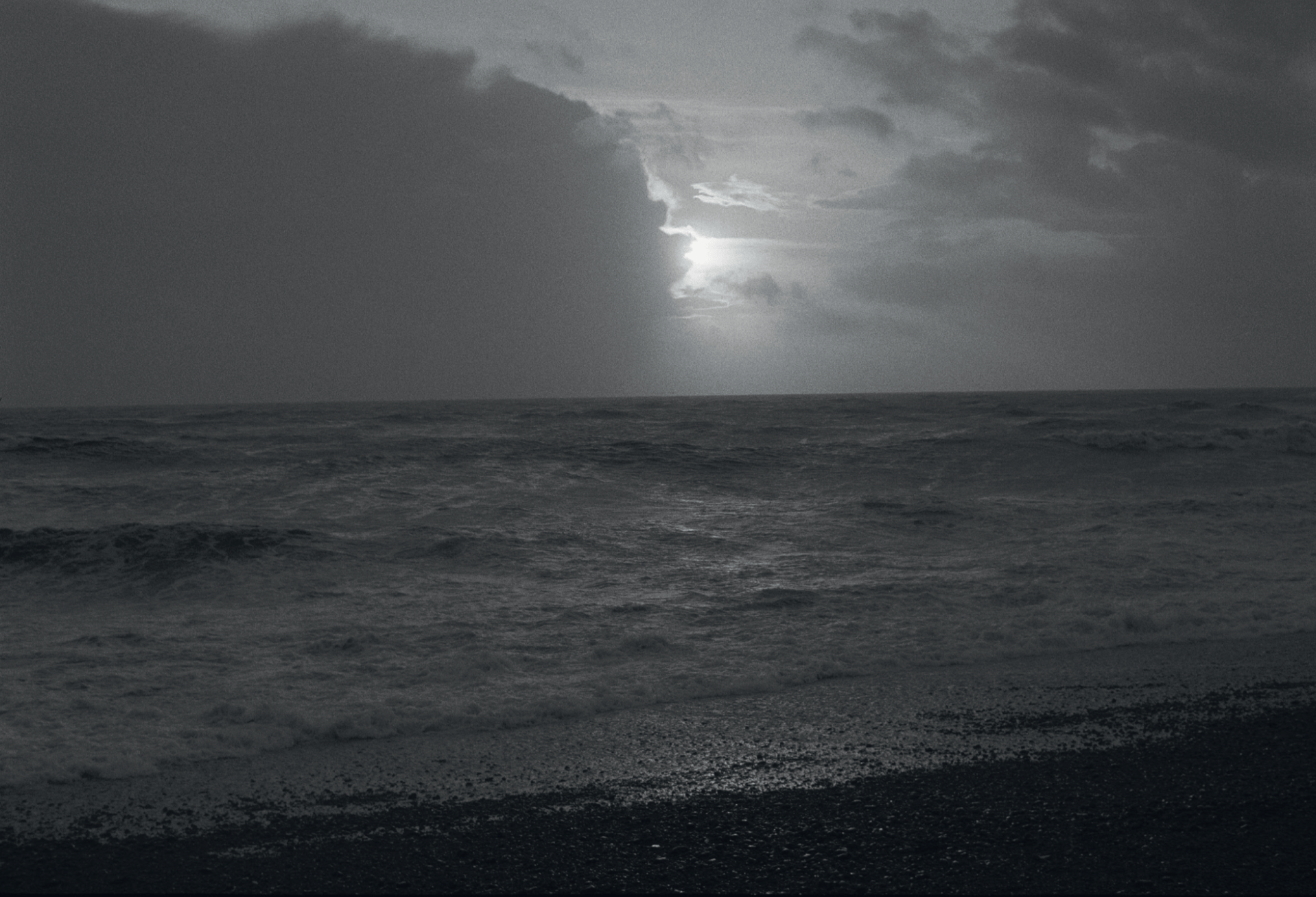 Black-and-white photo of the beach, the sunlight peering out from behind the clouds