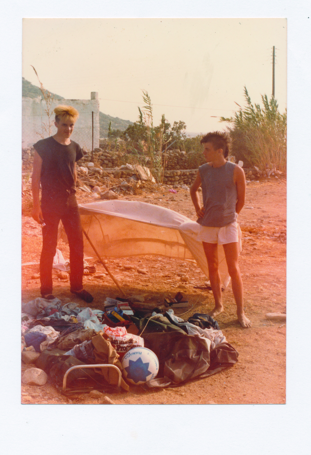 Photo of two people standing outside in the sand and heat next to a lateral shelter, clothes and a ball