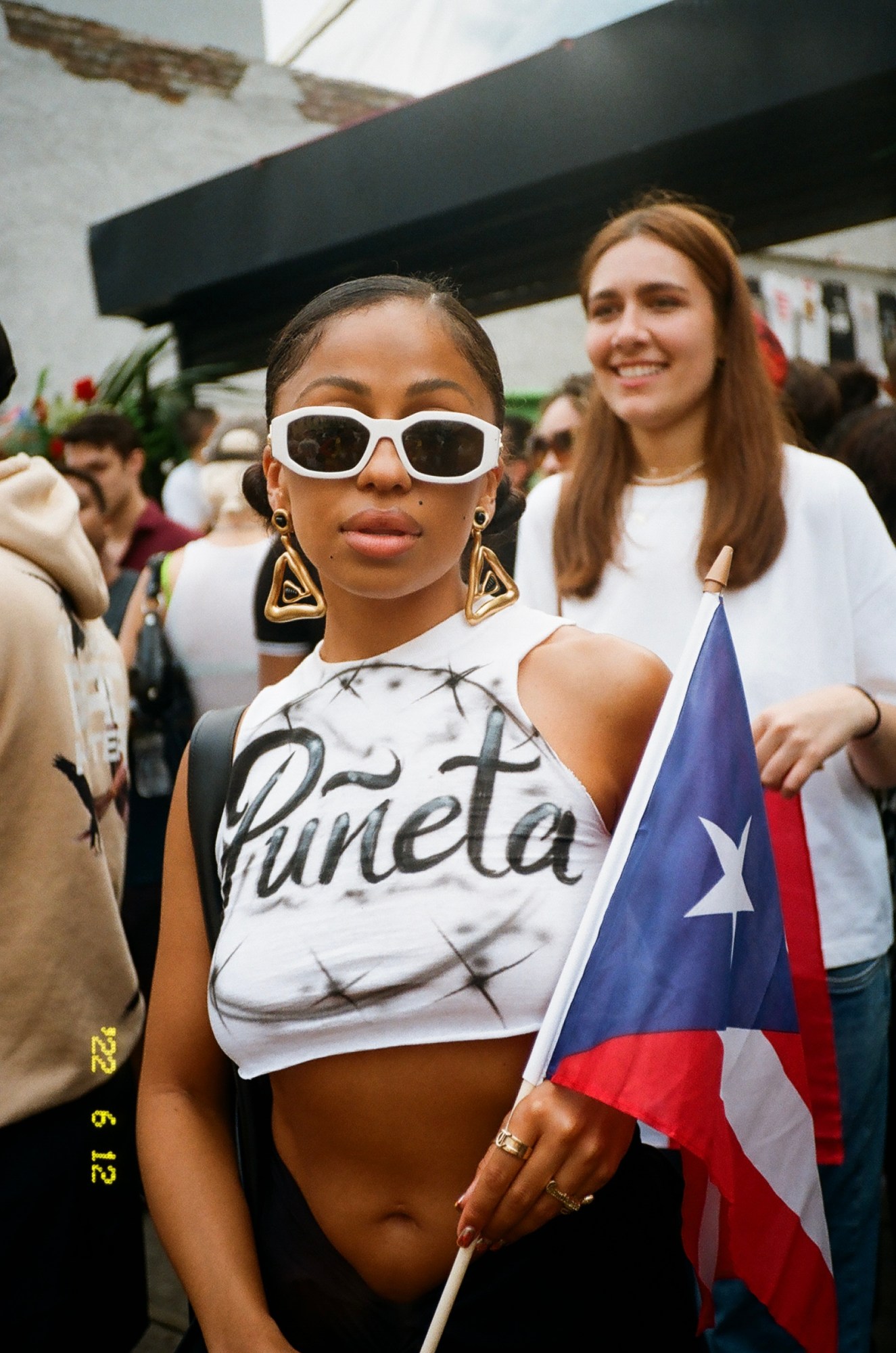 Woman in Bushwick wearing sunglasses, white Puñeta t-shirt, carrying Puerto Rican flag