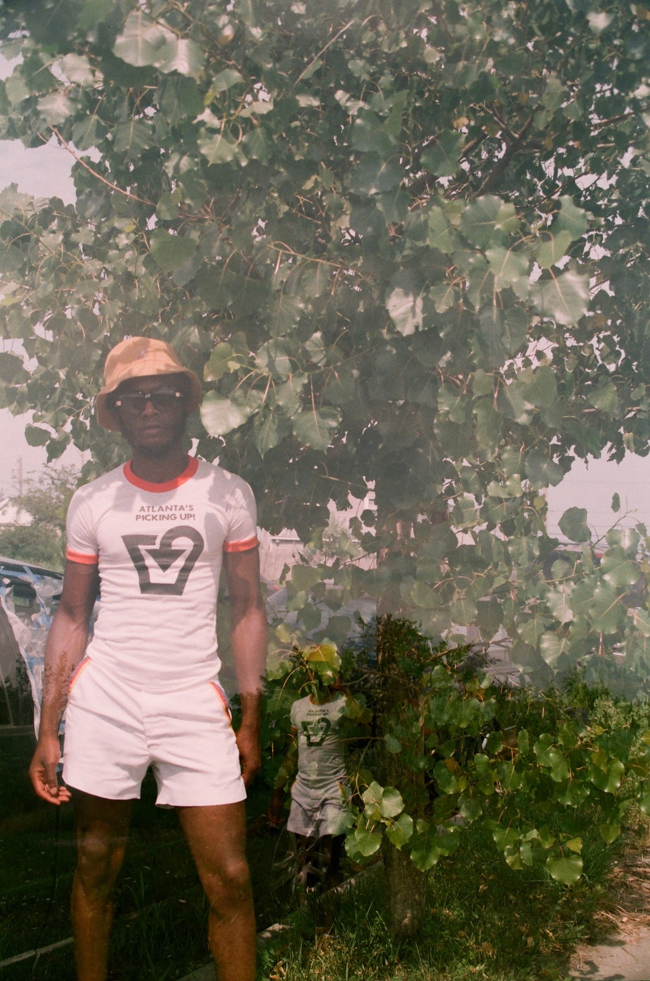 A man in white shorts and t-shirt standing in front of a bush in Queens