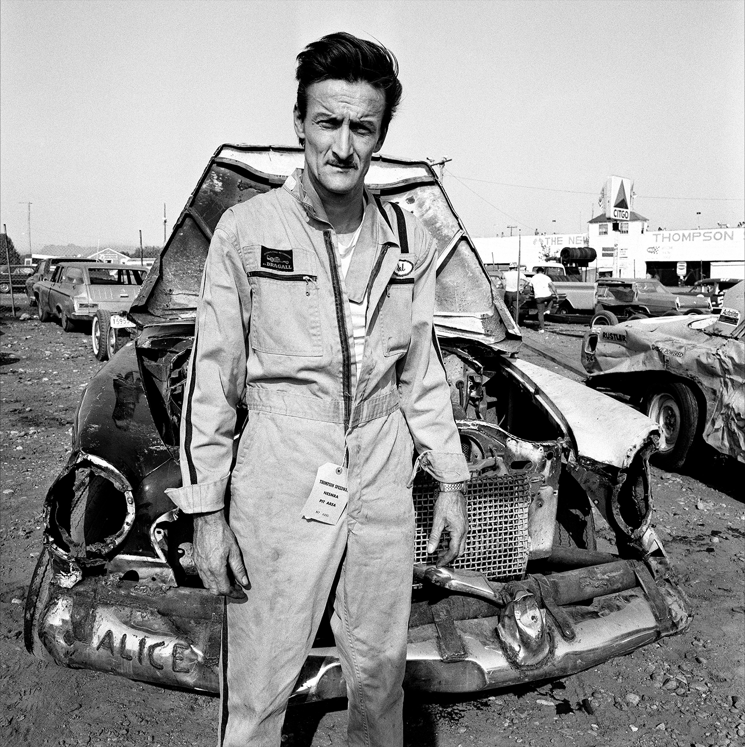 a man in an unzipped boiler suit stands in front of a wrecked vehicle named 'alice' according to its front bumper