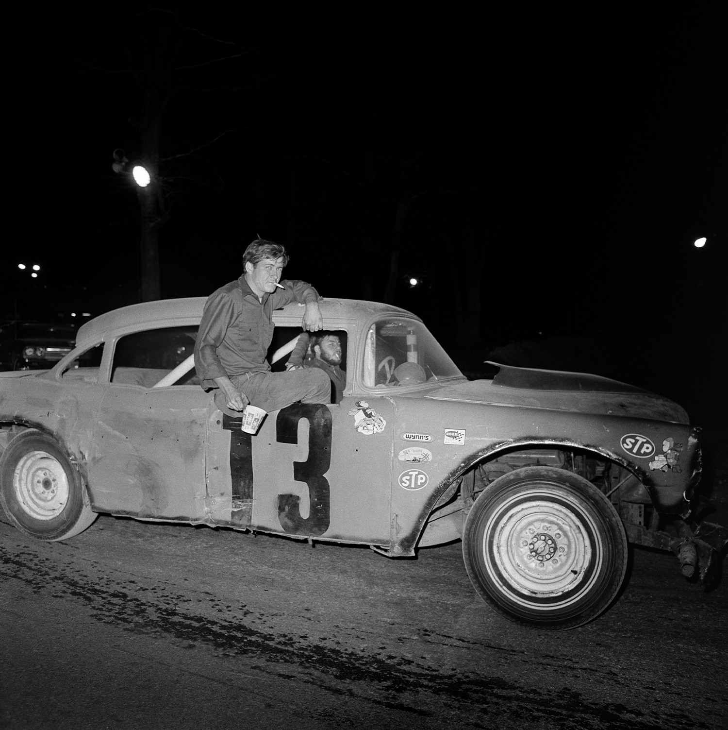 two men smoking cigarettes ride in a car marked with the number 13, one is driving and the other leaning out of the side window