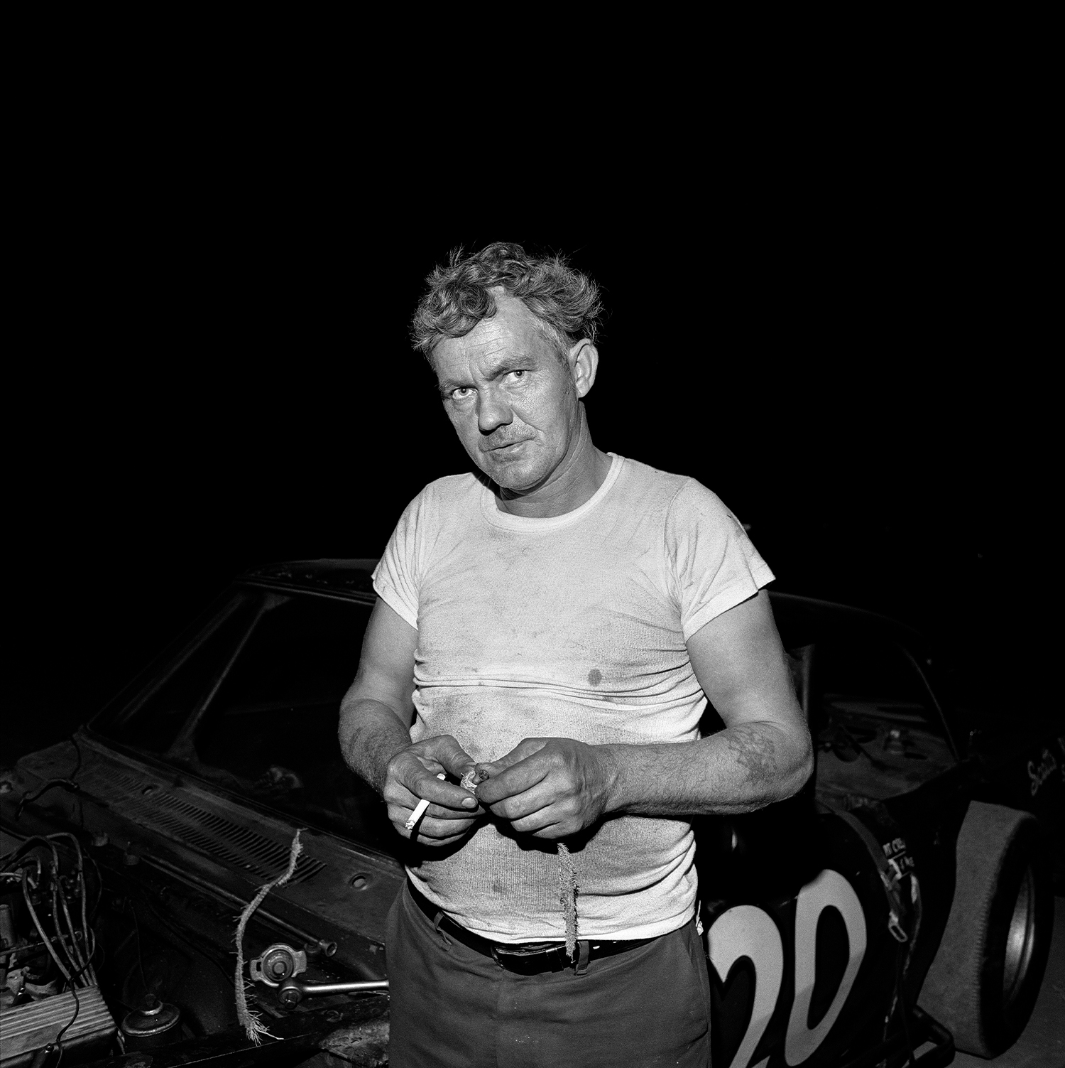 an older man, standing in front of a race car, looks off frame as he fiddles with something in his hands and smokes a cigarette