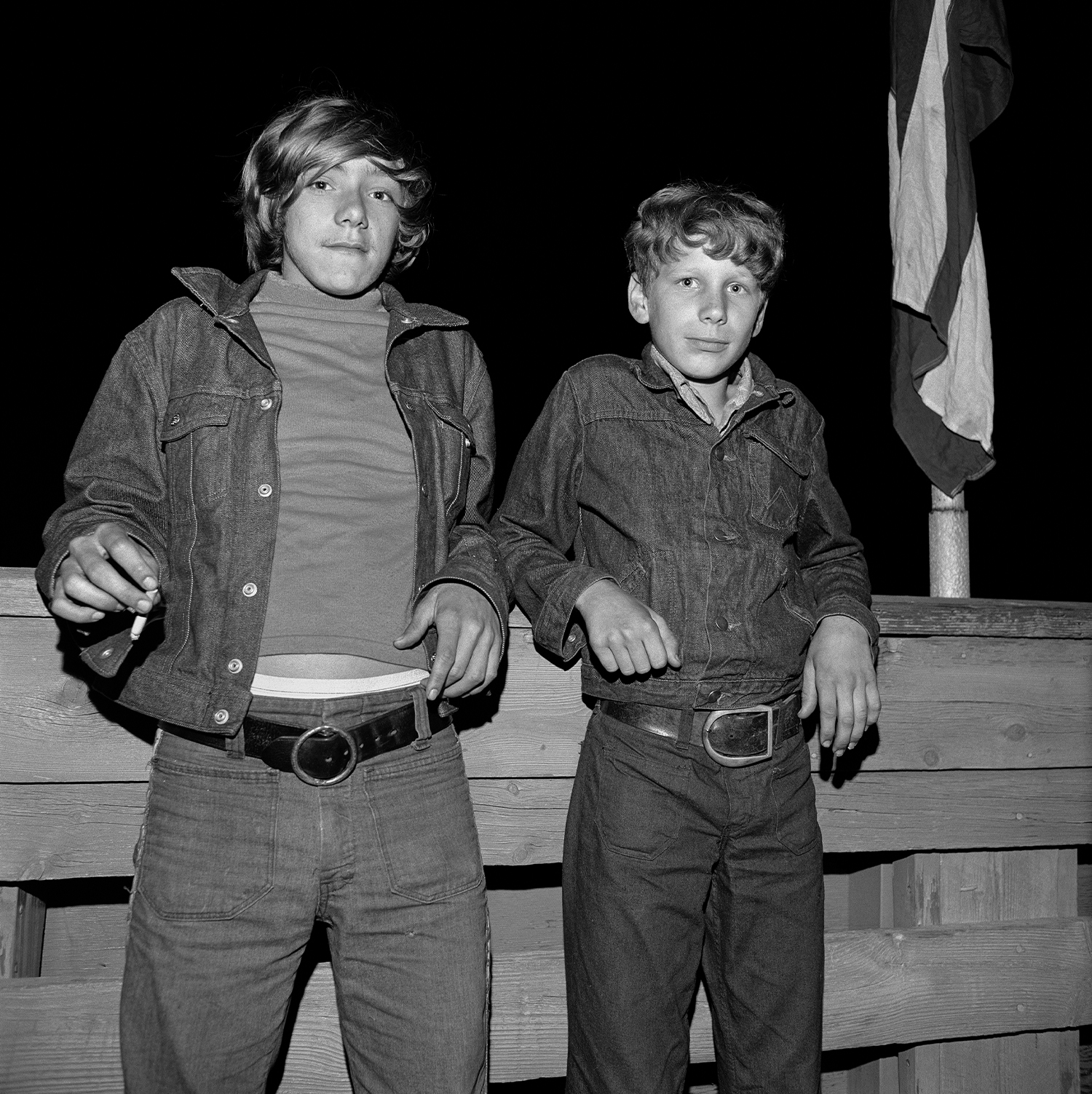 two young boys lean against a wooden structure, one smoking a cigarette