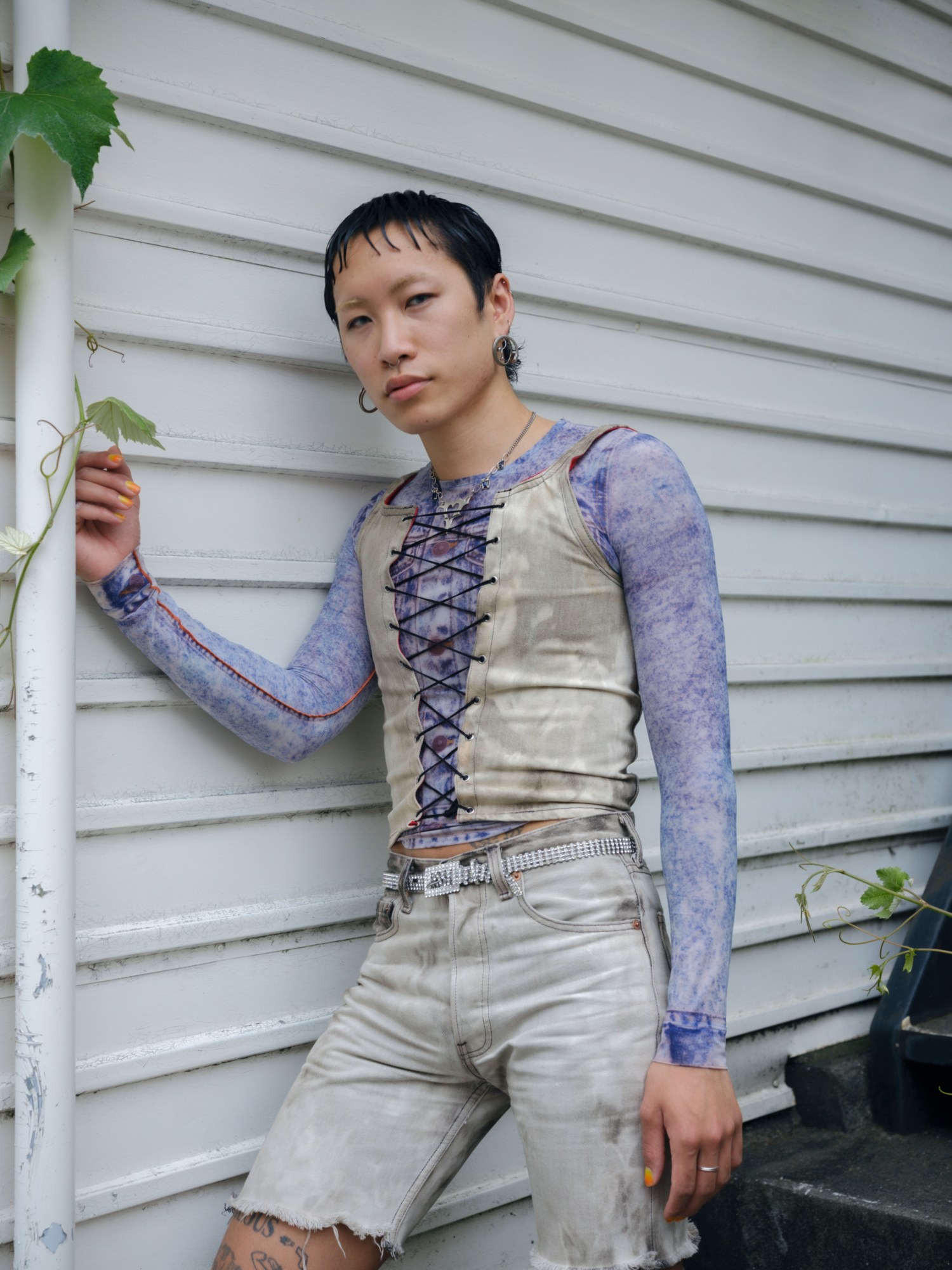 a person wearing denim shorts and a bodice style shirt stands leaning against the side of a panelled buidling