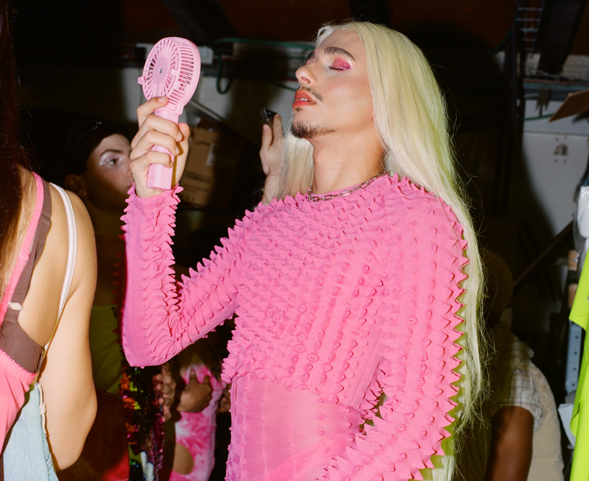 A young Orthodox Jewish fans his face while wearing a pink outfit and blond wig at a drag ball.