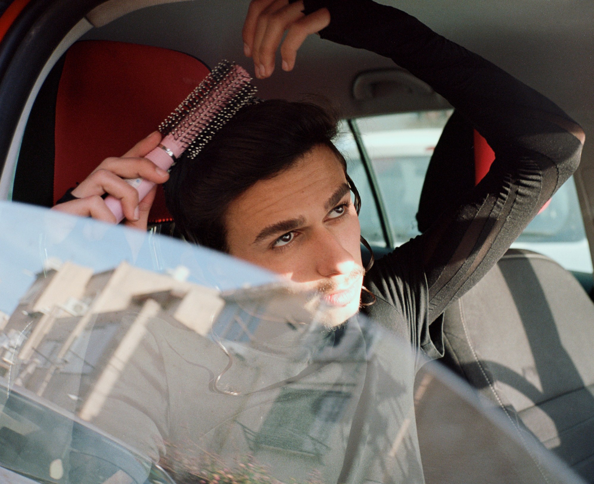 A young Orthodox Jewish combs his hair with a pink brush in the front seat of a car.