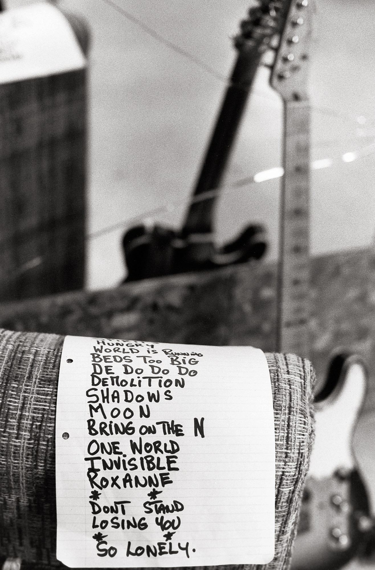 a handwritten set list rests over a cushion in a backstage room, a guitar stands out of focus in the background