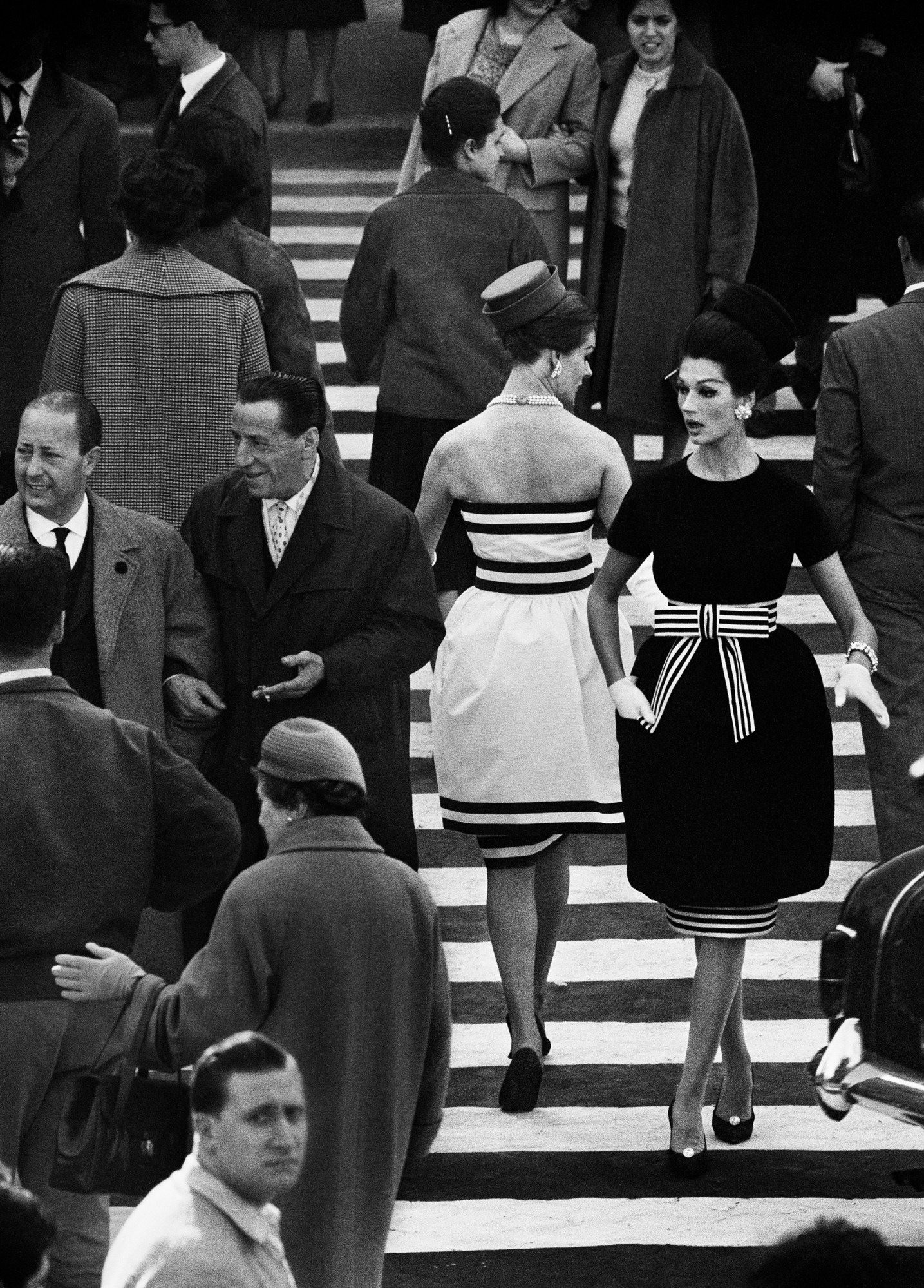 Black-and-white photo of people in suits and cocktail dresses walking along a striped road