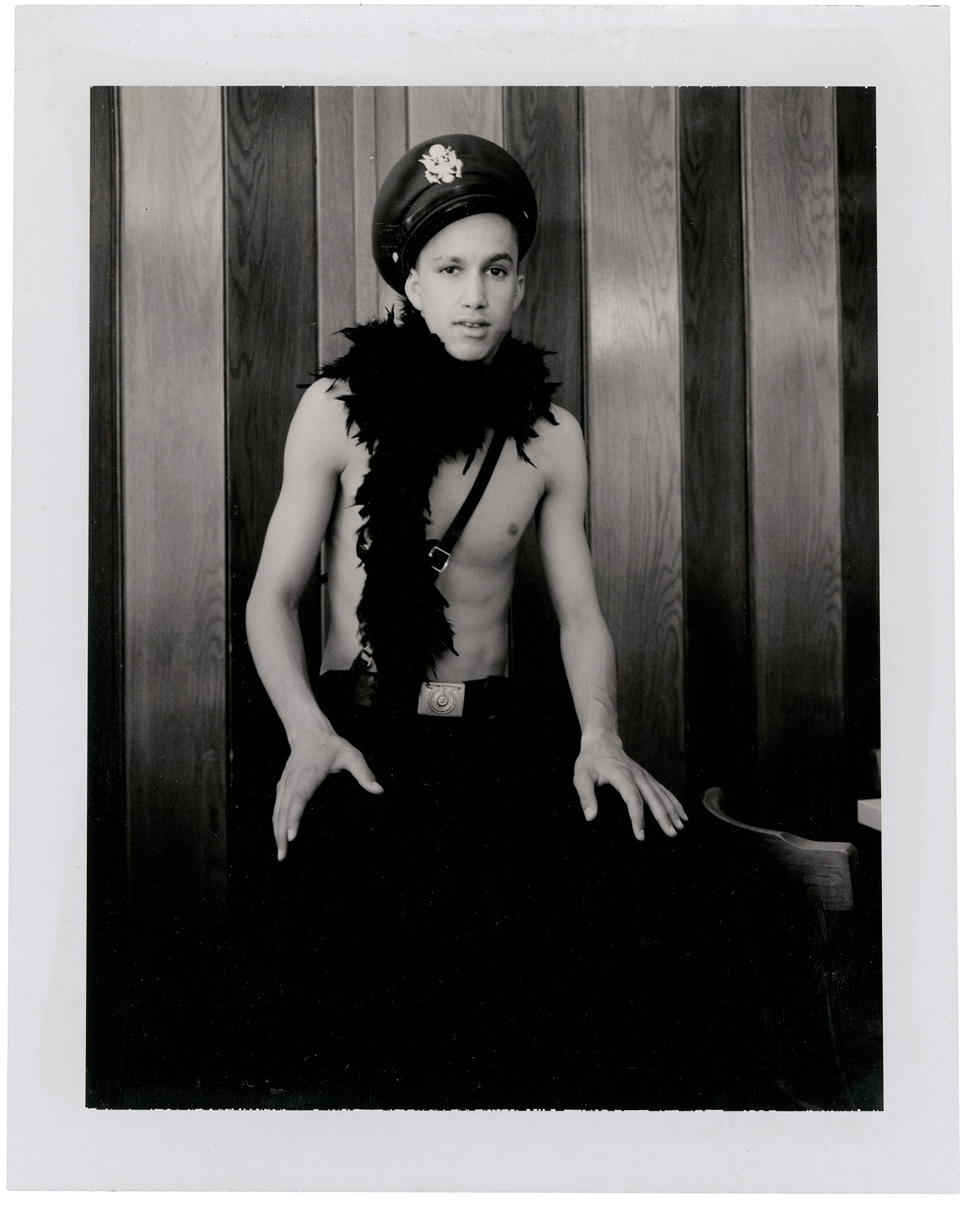 Polaroid of a boy in German military trousers, belt and hat and a feather boa sitting in a barn.