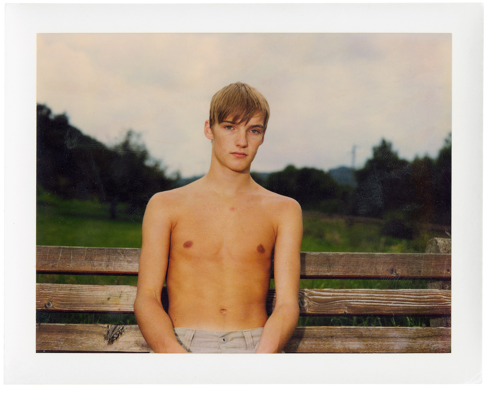 Polaroid of a shirtless boy in denim trousers sitting on a bench outside in a park