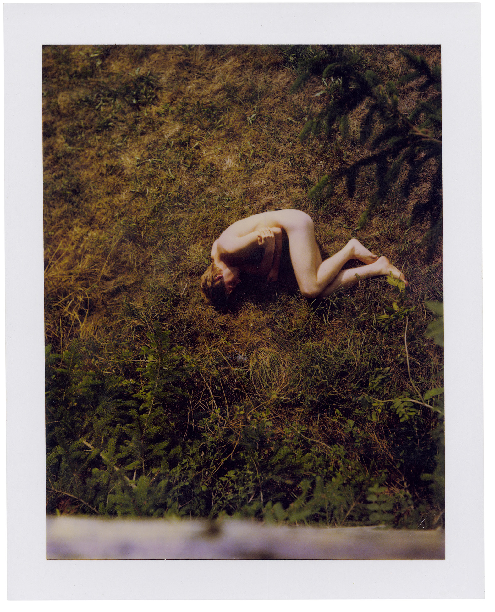 Polaroid of a naked boy holding himself while lying on the grass.