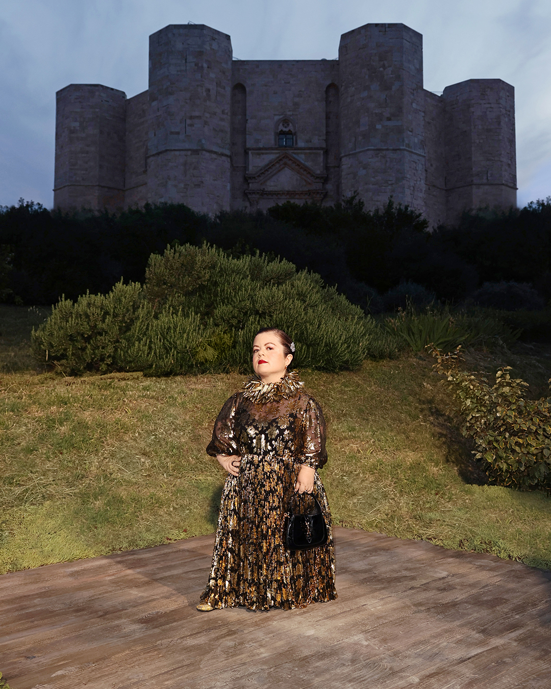 An image of Sinéad Burke wearing a gold lamé gown, stood against the ba