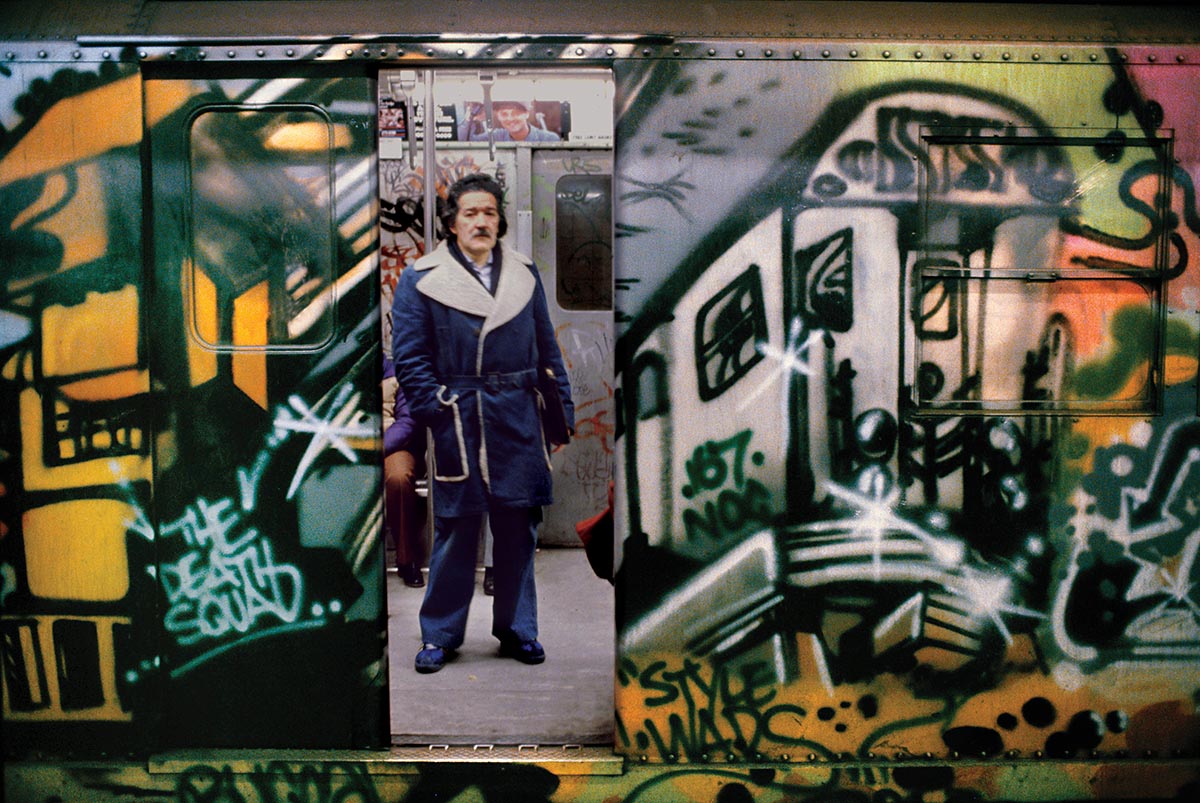 a man with a moustache stands in an open doorway of a metro carriage covered in colourful graffiti