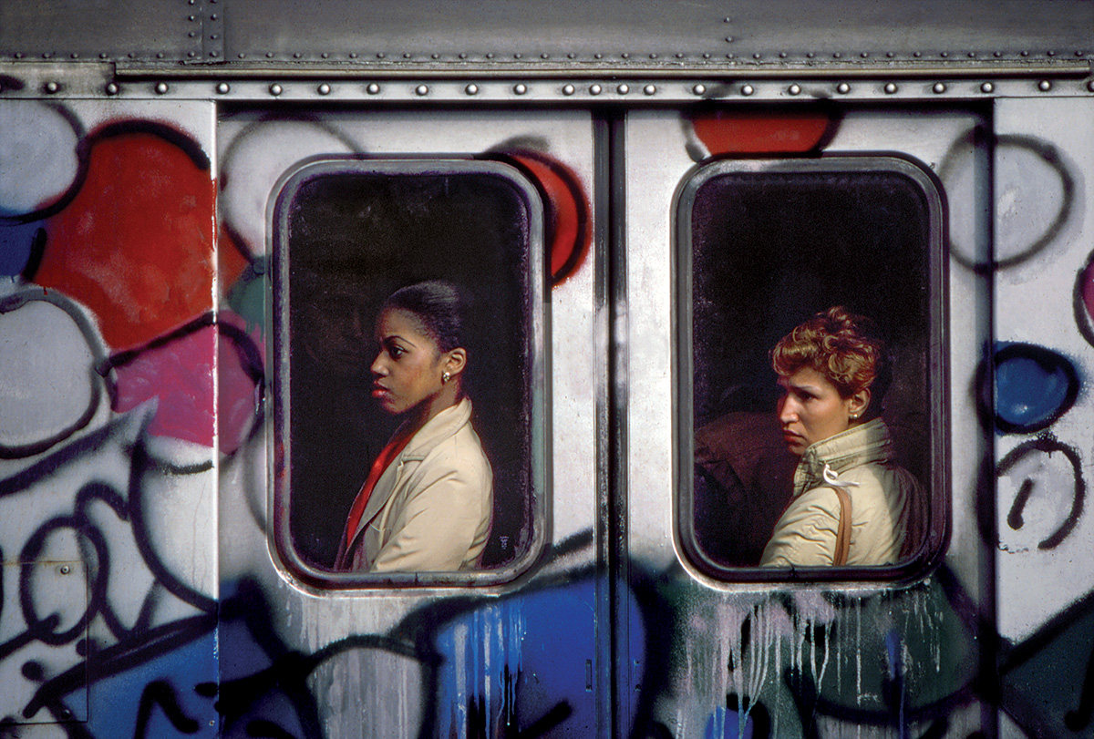 two women in beige trench coats peer out of the windows of a graffiti-covered subway car