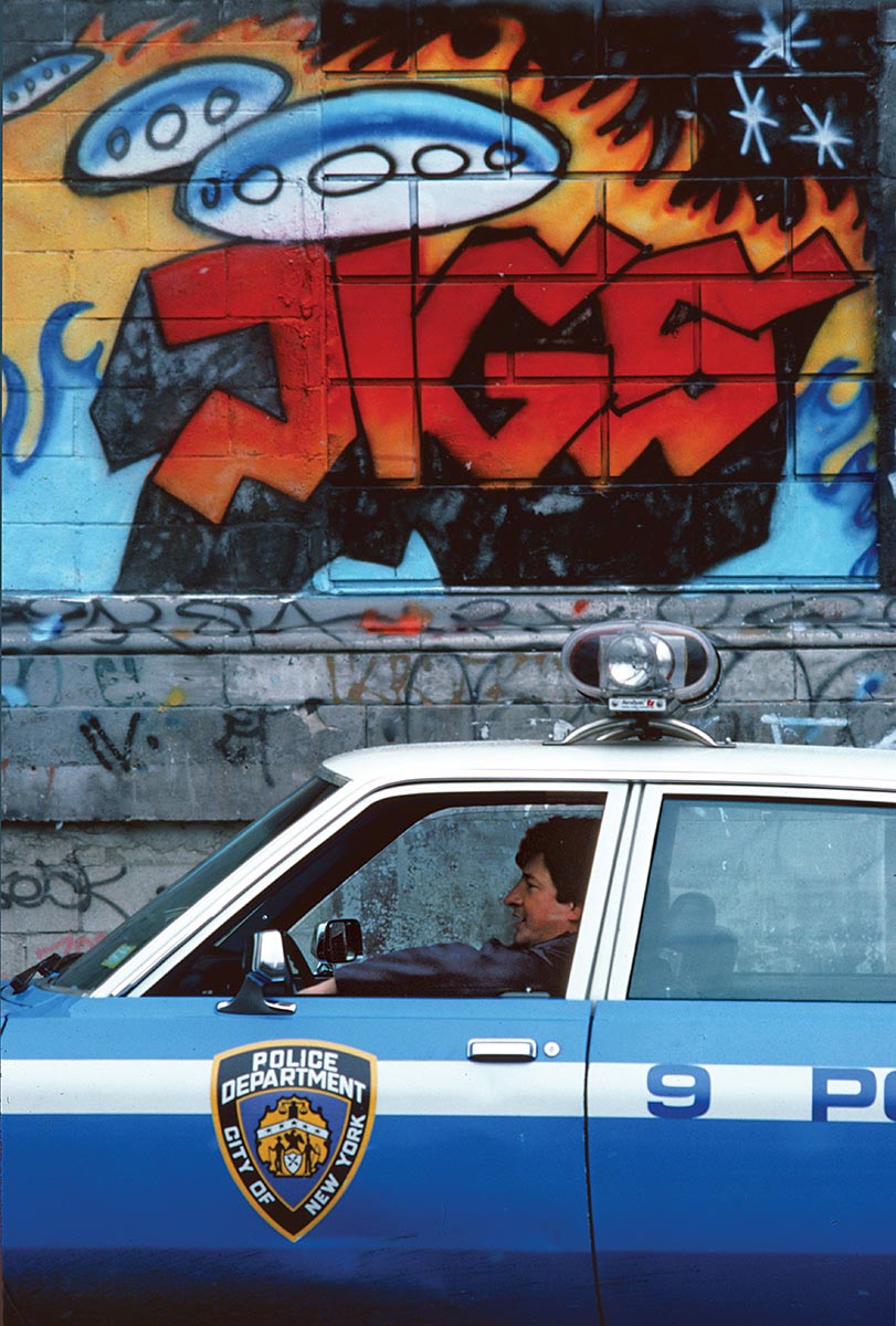 a policeman sits in an 80s-style NY cop car in front of a graffitied wall