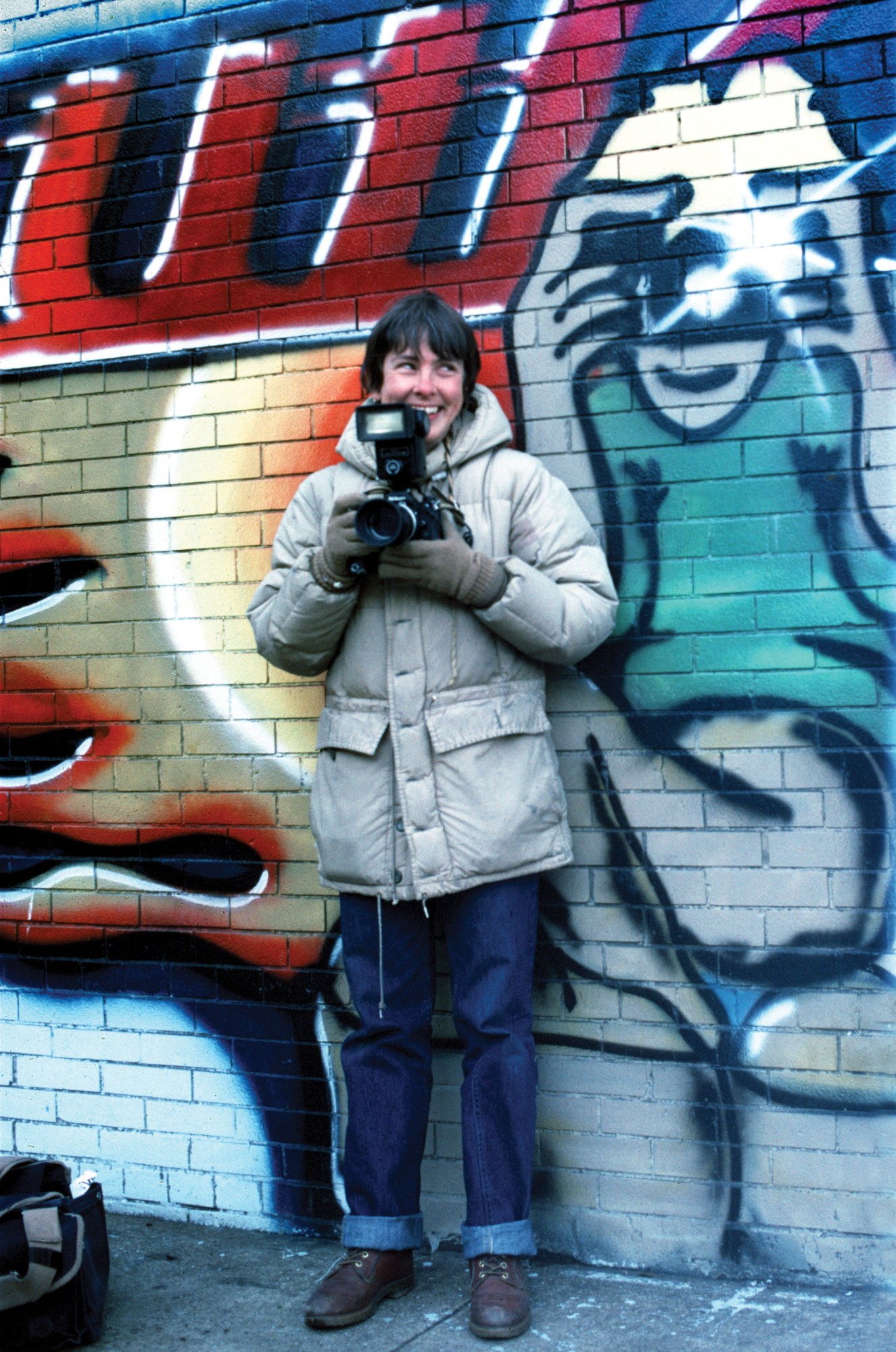 the photographer Martha Cooper wrapped up warm and holding her camera, in front of a wall of graffiti