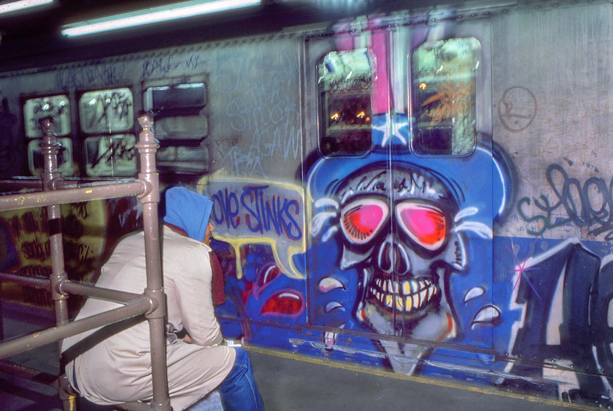 a person sits on a train platform, in front of a graffitied skull and the painted words: LOVE STINKS