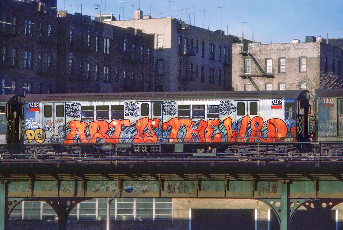 a subway car with the phrase ART IS THE WORD spray painted on it goes over a high line in NYC